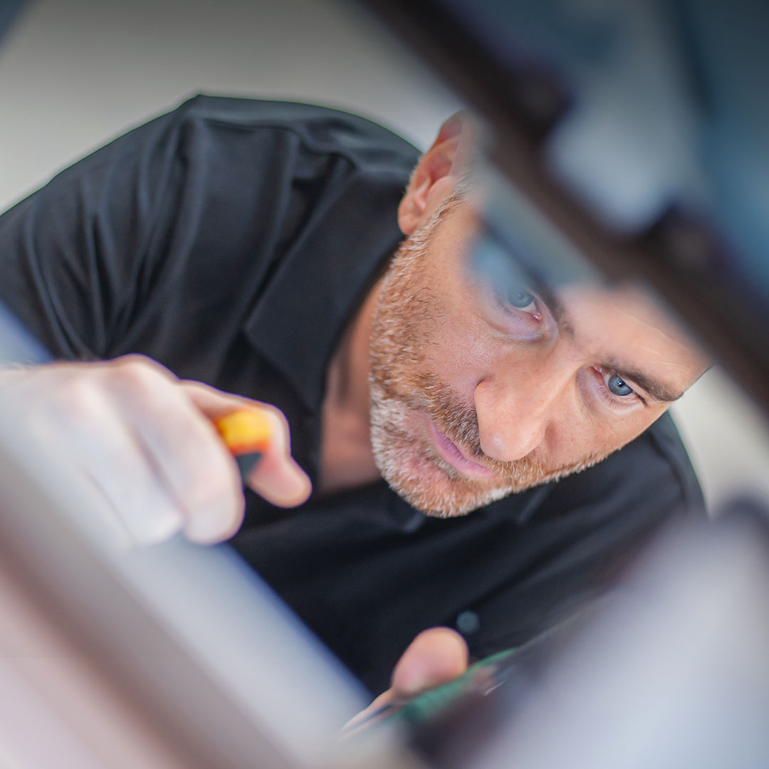 Bearded man in black shirt concentrates on repairing or assembling something with a tool, blurred foreground frames his focused face.