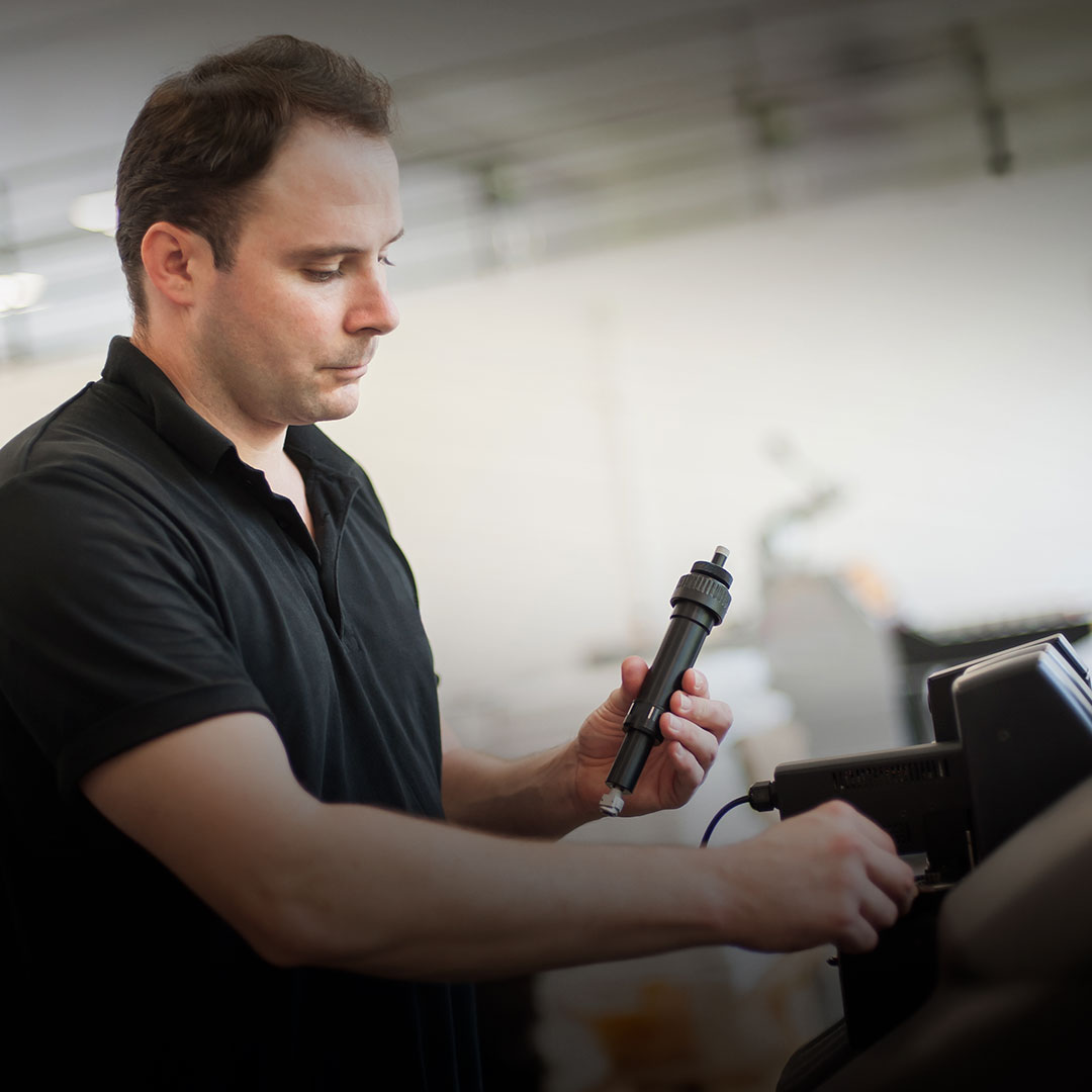 Man in black shirt operates handheld power tool on equipment inside a well-lit indoor workshop.