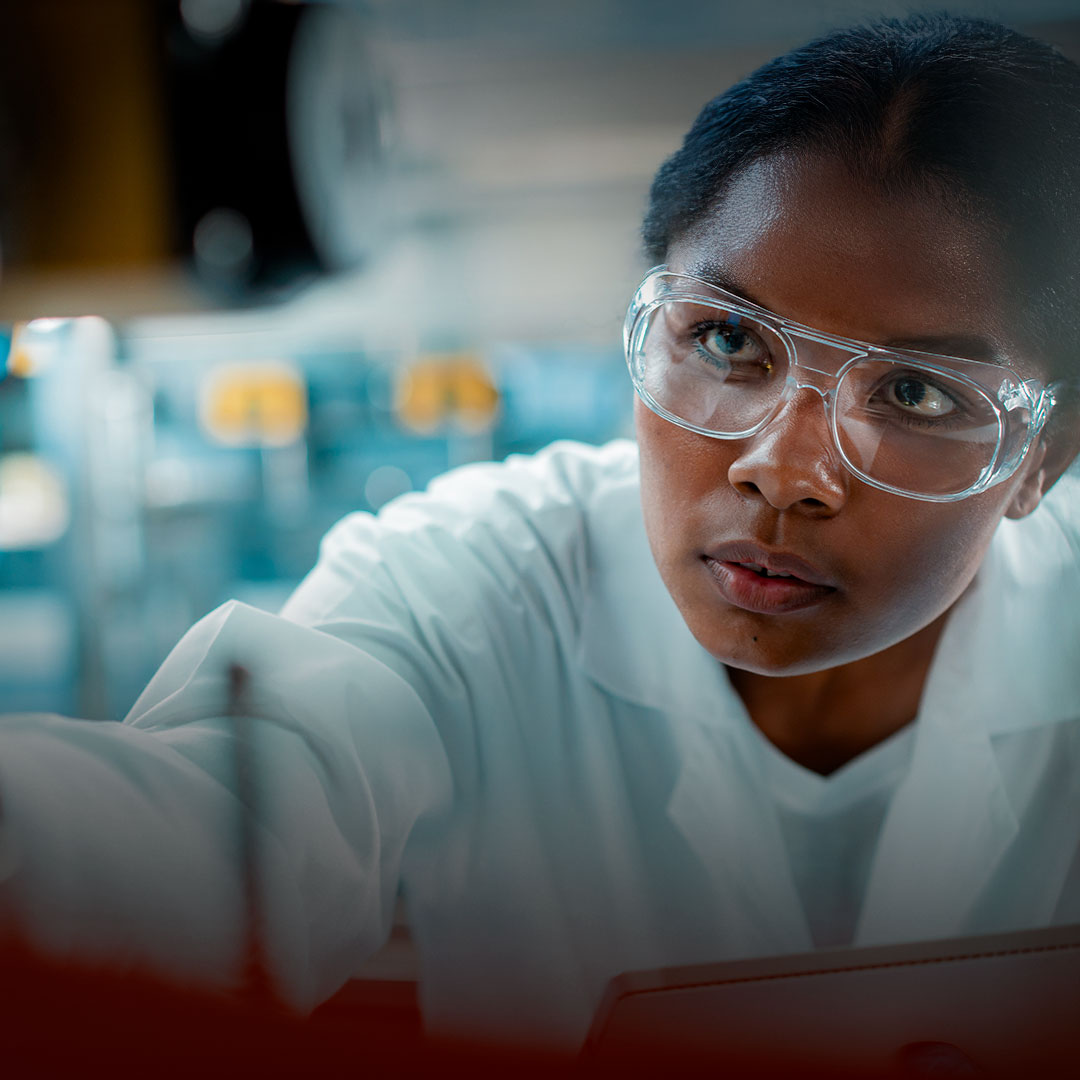 Female scientist in lab coat and safety goggles examining equipment, blurred lab background emphasizes her focus and scientific setting.