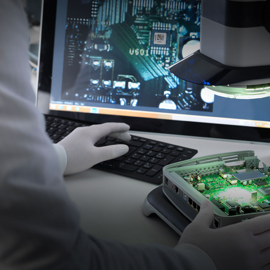 Person in white gloves inspects circuit board in open device using magnifier and keyboard, monitor shows detailed circuit diagram.