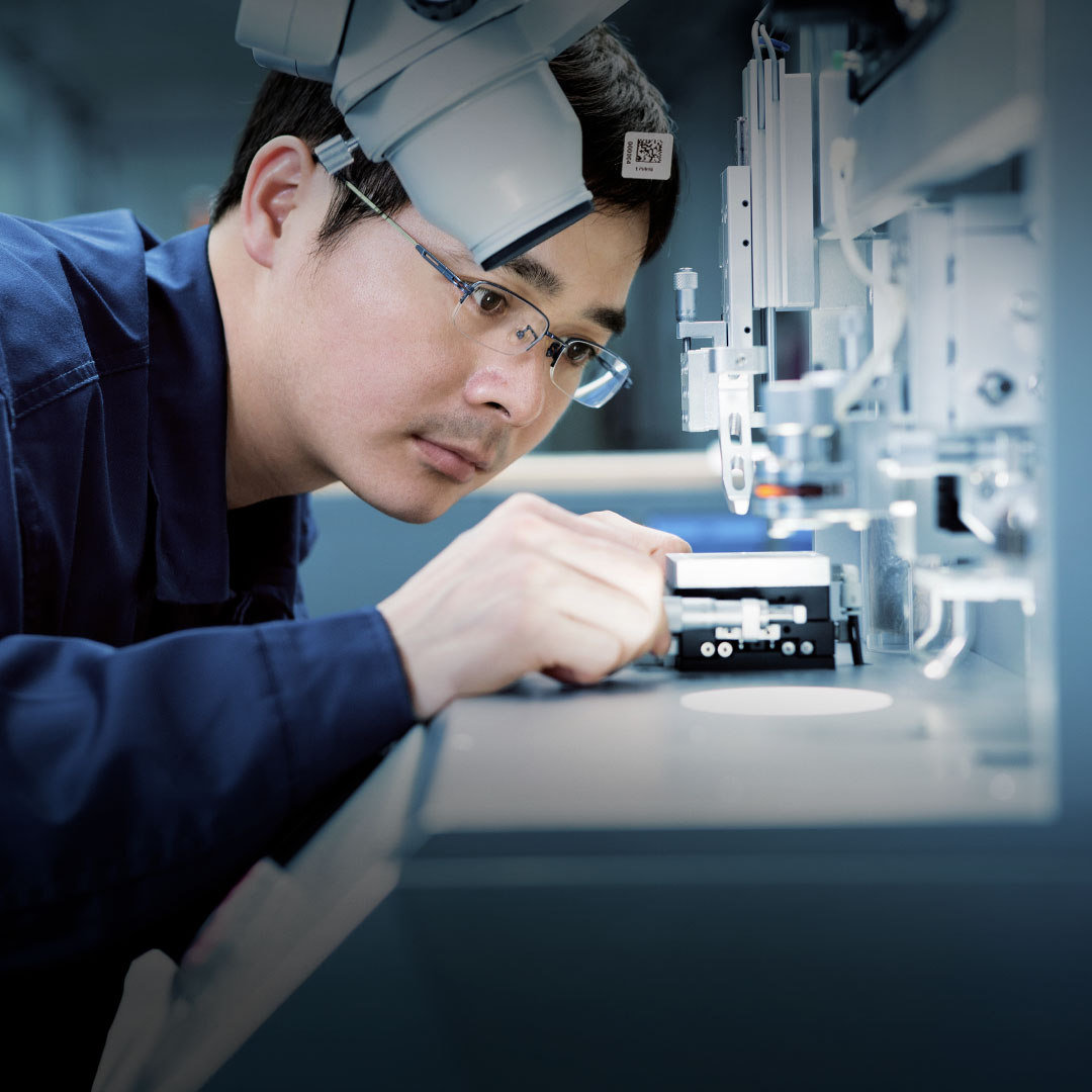 Man in glasses and blue lab coat examines small device under microscope in a modern laboratory or high-tech manufacturing setting.