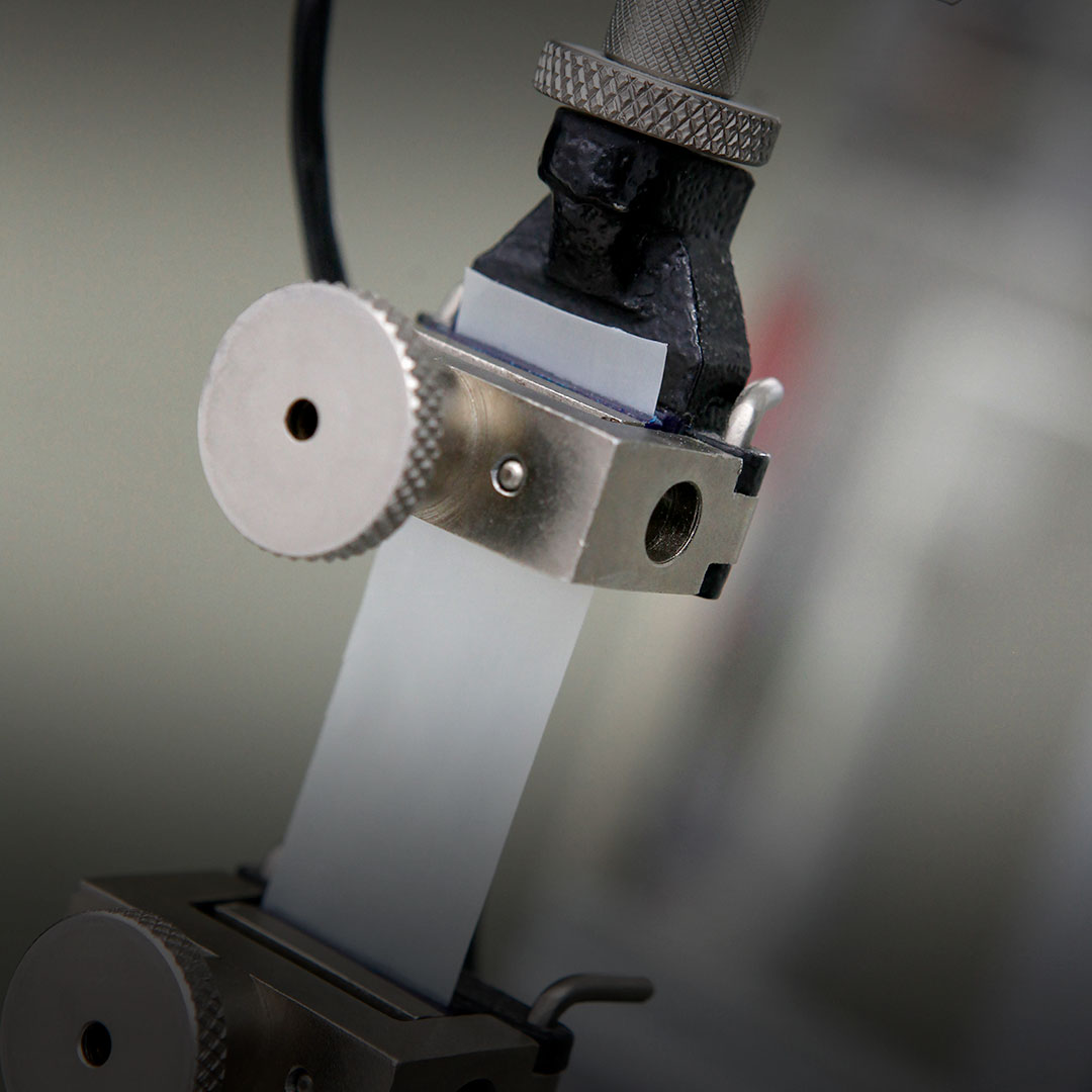 Mechanical device clamps a translucent rectangular strip, featuring metallic knobs and textured surfaces in a lab setting.