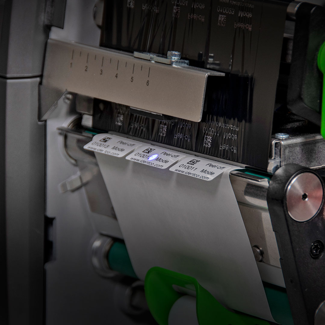 Label printer printing barcode labels on a white strip, featuring metallic and green parts and a blue light highlighting one label.
