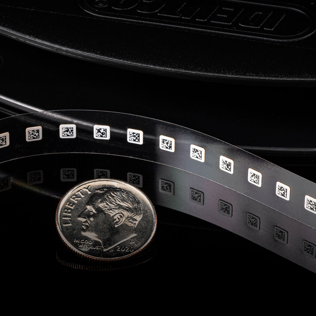 U.S. dime beside a row of tiny QR code labels on clear film, highlighting the small label size compared to the coin.