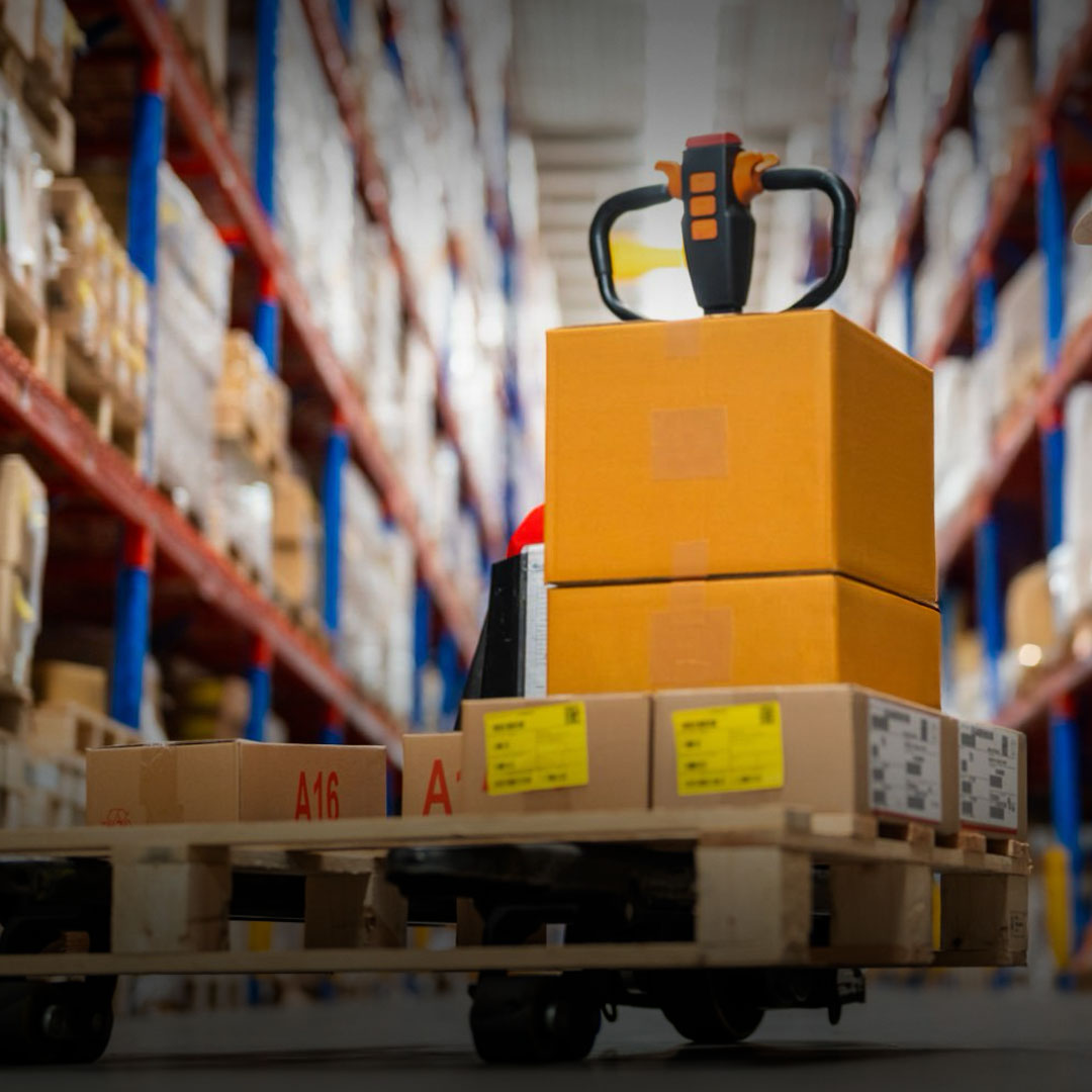 Pallet jack with cardboard boxes in a warehouse aisle amid tall shelves stocked with packages and inventory.