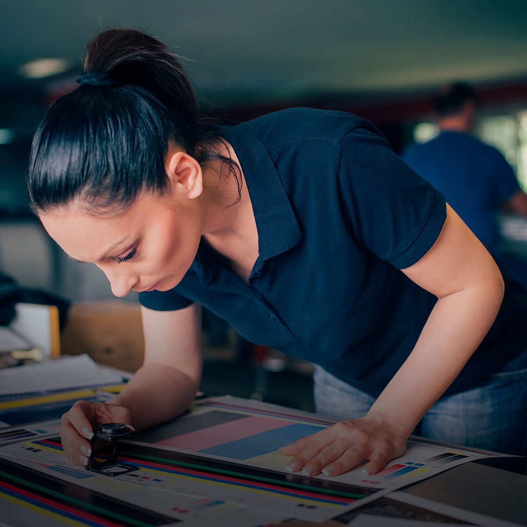 Woman in navy polo shirt with dark ponytail inspects color print samples on table using magnifying glass to check print quality.