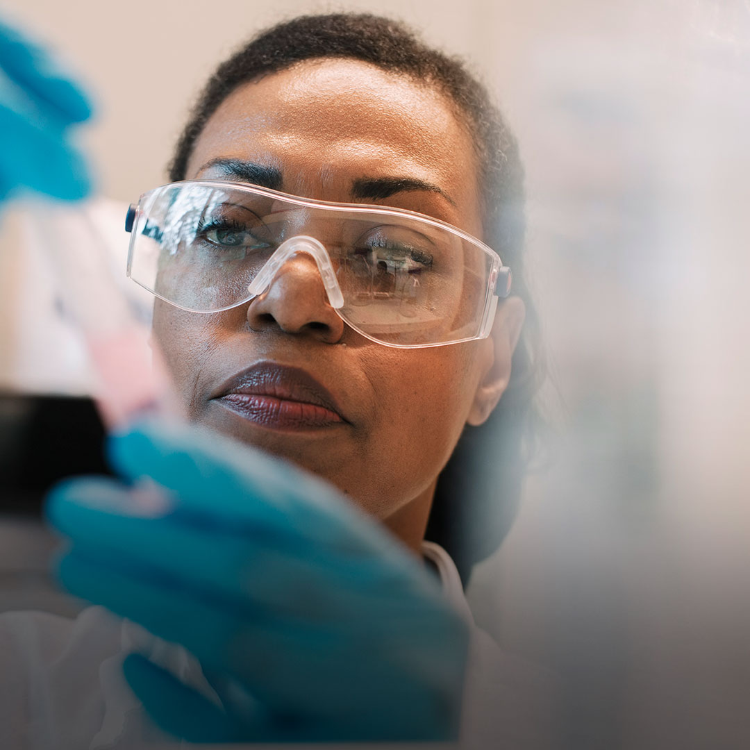 Scientist in safety goggles and blue gloves studies a test tube of pink liquid with concentration in a laboratory setting.