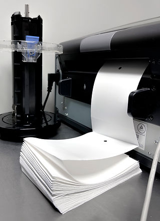 Freshly printed white paper stacking in a desktop printer, with a black cylinder and small blue container beside it.