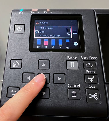 Finger pressing button on printer control panel with color screen showing paused status, material type, ink levels, and function buttons.