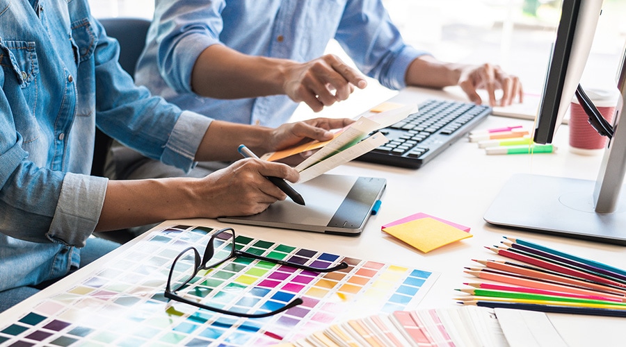 Designers collaborate at a desk with color swatches, colored pencils, tablet, computer, glasses, and sticky notes.