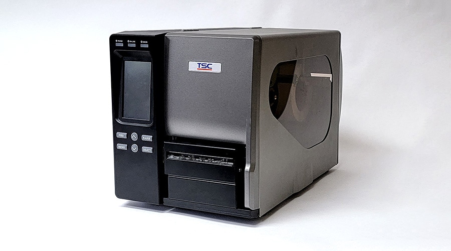 Gray industrial label printer with display, control buttons, and transparent side panel showing label roll on white background.