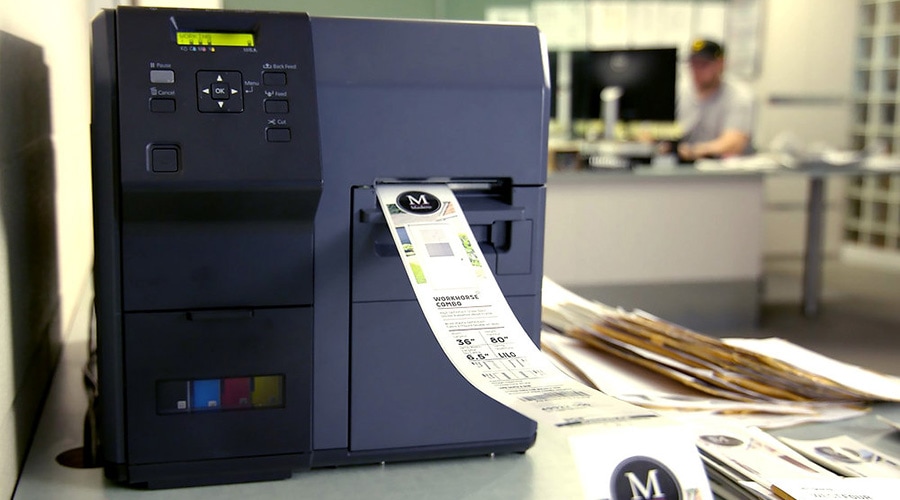 Label printer printing a shipping label in an office, with papers and a person working at a desk blurred in the background.