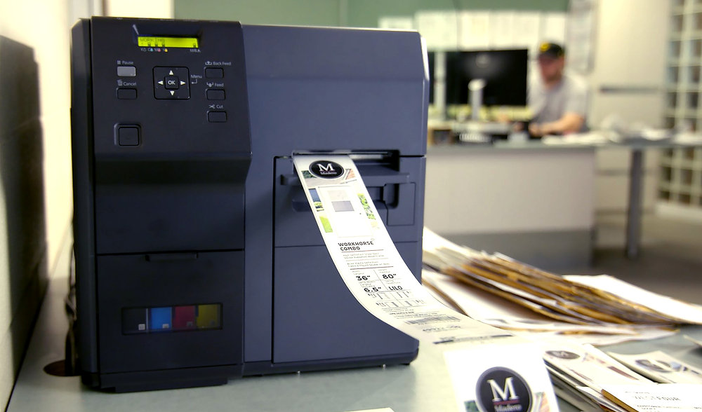Label printer producing a color product label on a desk with scattered papers; person works on computer in blurred office background.
