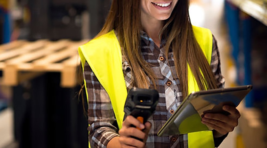 Smiling woman in a safety vest and plaid shirt holds a barcode scanner and tablet in a warehouse environment.