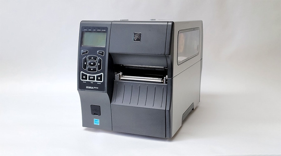 Zebra industrial label printer with left display and controls, central label slot, clear side panel, on a plain white background.