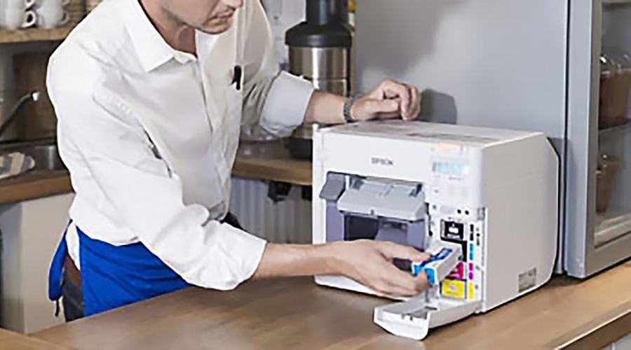 Person in white shirt changes ink cartridge in Epson printer on wooden counter in kitchen or office workspace.