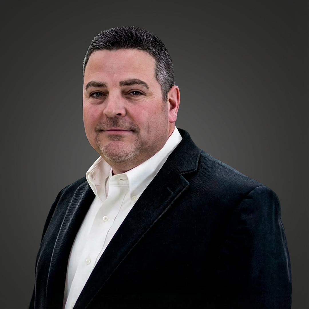 Confident man with short dark hair and trimmed beard in white shirt and dark blazer stands against a plain dark background.