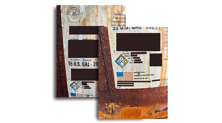 Rusty metal barrels with faded, stained labels featuring a U.S. flag and hazard symbol; some label info blocked by black boxes.