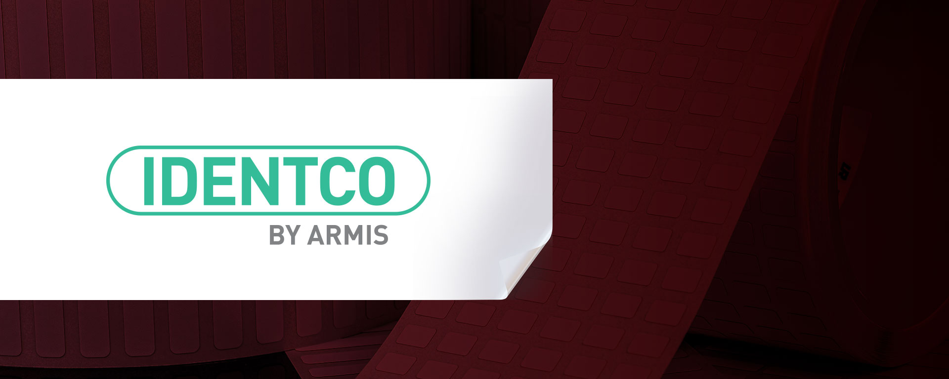 White sticker featuring green and gray IDENTCO BY ARMIS logo, partially peeled at corner on dark red grid-patterned background.