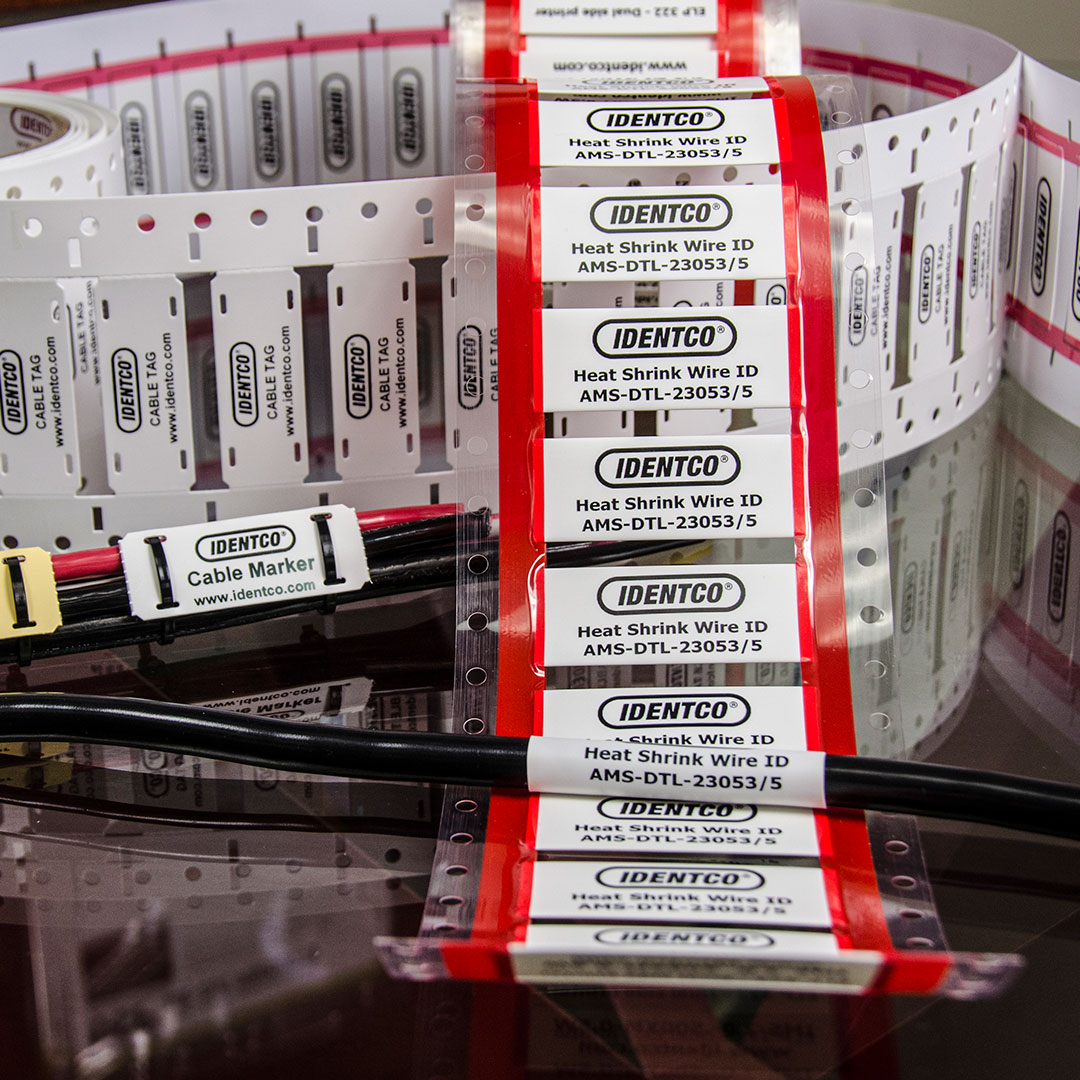 IDENTCO red and white heat shrink wire ID labels on black cables, with extra rolls in background; model AMS-DTL-23053/5 shown.