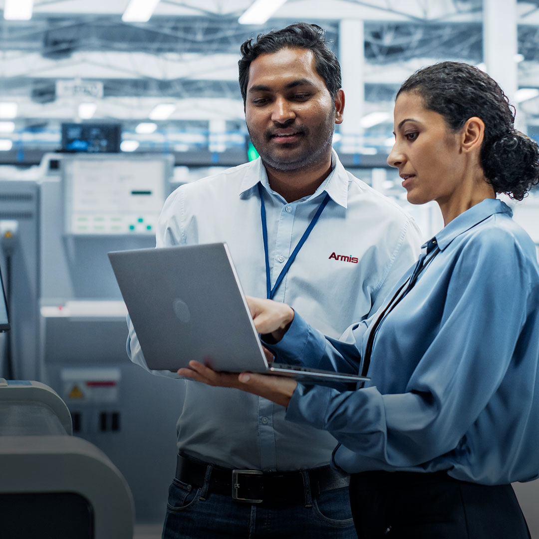 Two professionals in business attire review data on a laptop in a modern industrial facility with large technical equipment behind them.