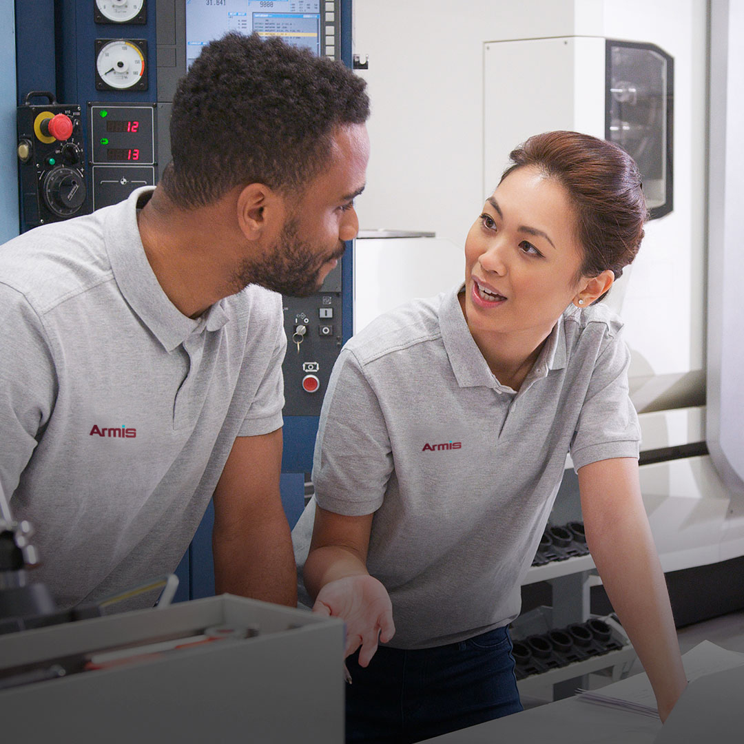 Two coworkers in gray Armis shirts talk near industrial equipment; the woman gestures as she speaks, and the man listens closely.