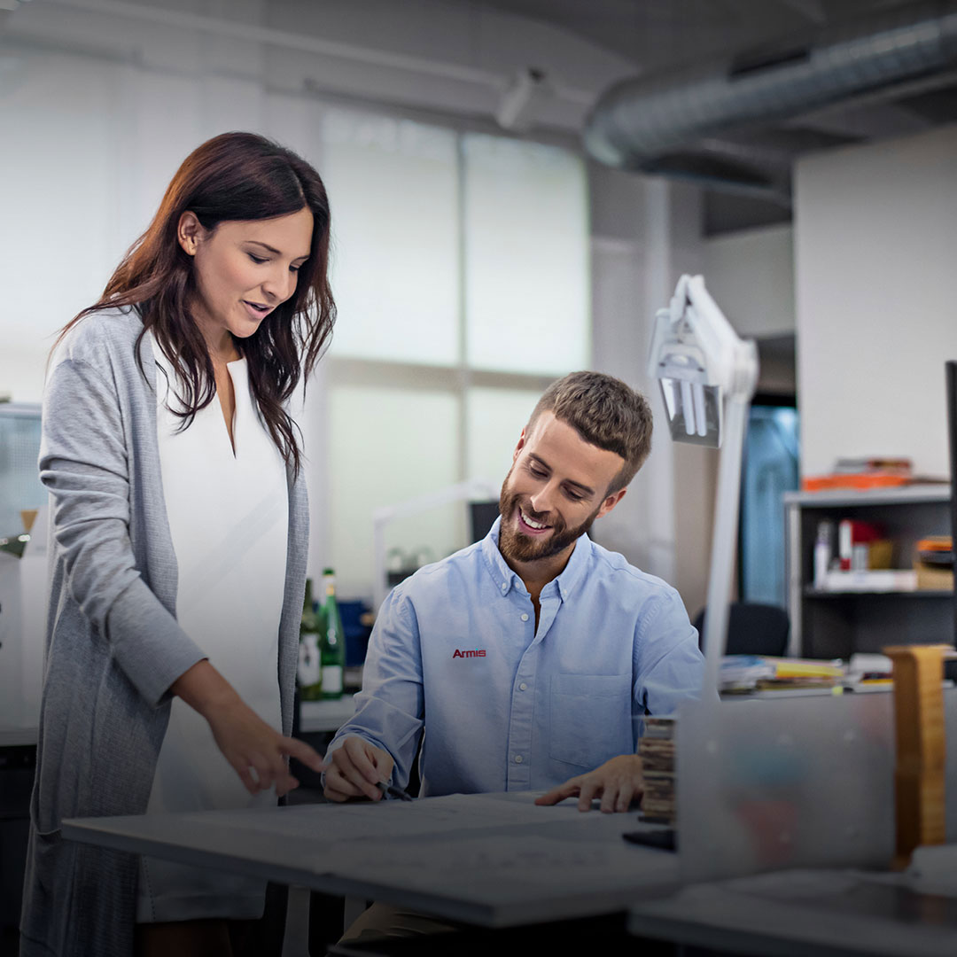 Woman stands by seated man at desk in modern office, both smiling at documents. Shelves and office equipment in background.