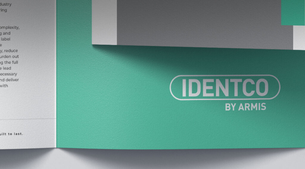 Green and white brochure titled IDENTCO BY ARMIS in bold, with partial printed page and small text visible on the left side.