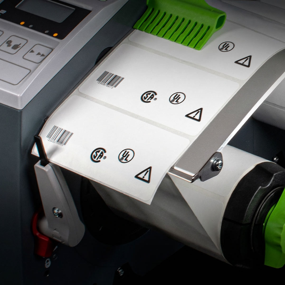 Label printer with digital display and green accents printing white labels featuring UL, barcode, and safety certification symbols.