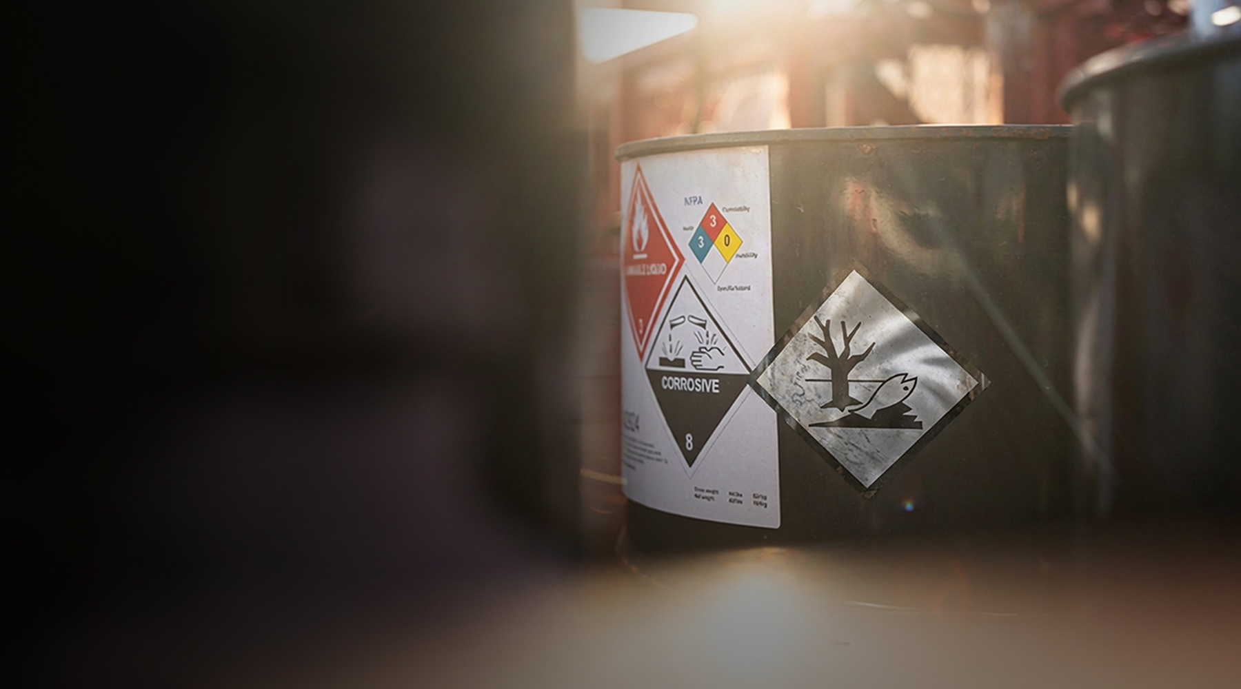 Green metal barrel with corrosive and environmental hazard warning labels in sunlight, blurred background.