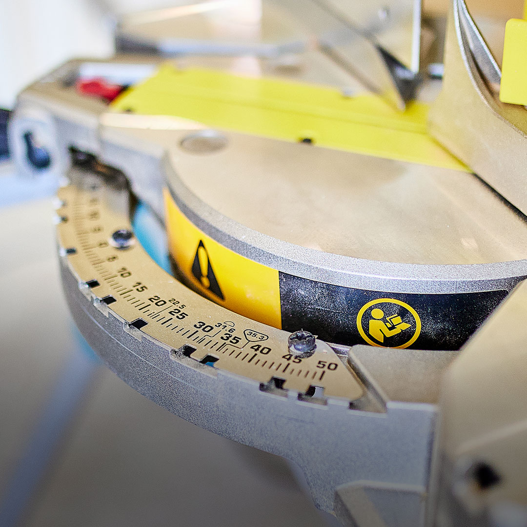 Circular saw angle gauge close-up with measurement markings, yellow caution label, and safety symbol warning to keep hands clear.
