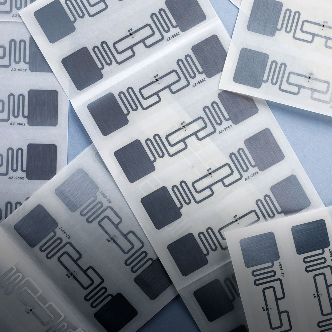 Sheets of adhesive RFID tags with metallic geometric circuits overlap on a blue background, showing electronic chip patterns.
