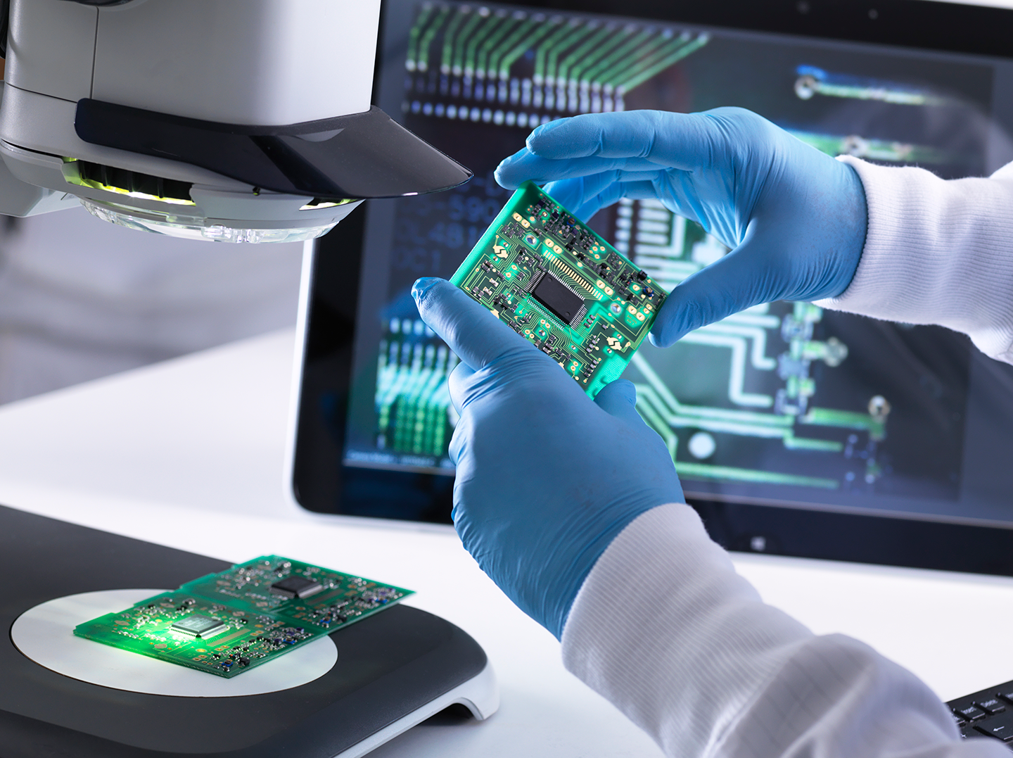 Technician in blue gloves inspects green circuit board under microscope with another board and computer showing circuit diagram nearby.