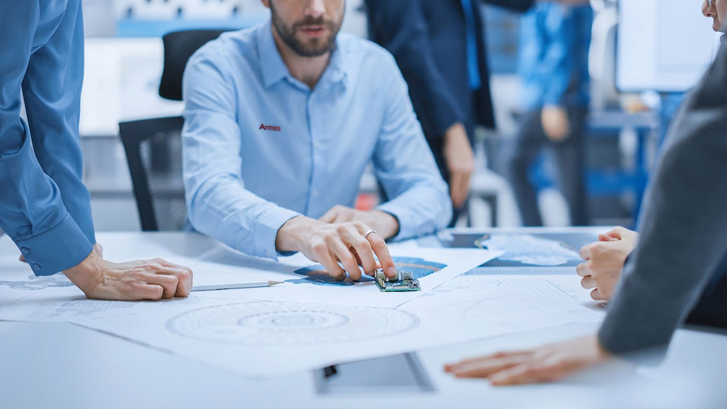 Business professionals review blueprints and a circuit board together at a table during an engineering design meeting.