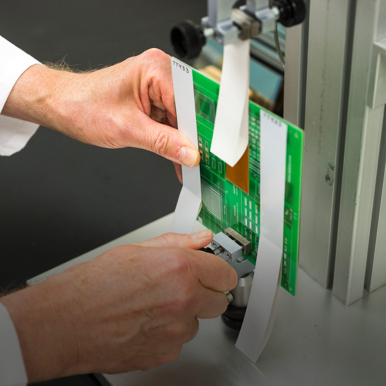 Person secures green circuit board with white paper strips in metal testing device for lab inspection or measurement.