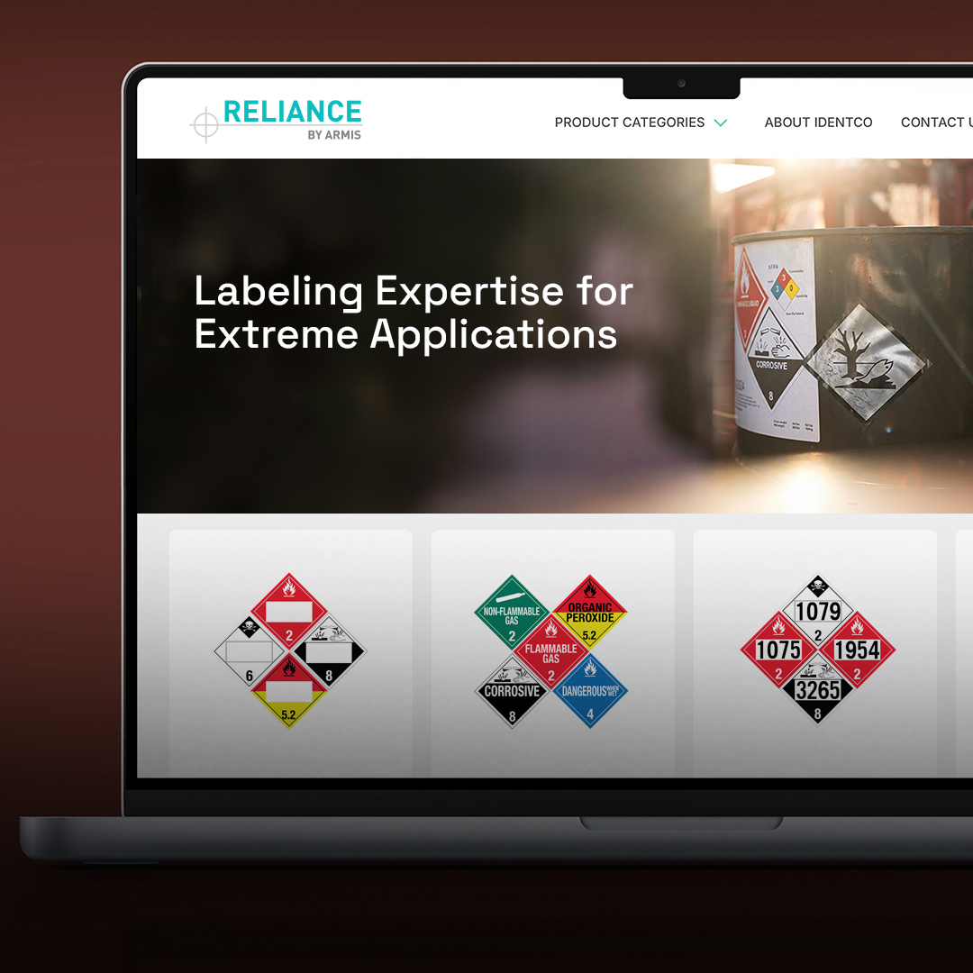 Laptop showing Reliance by Armis website with hazardous material label samples, warning symbols, and drum photo; menu visible top.