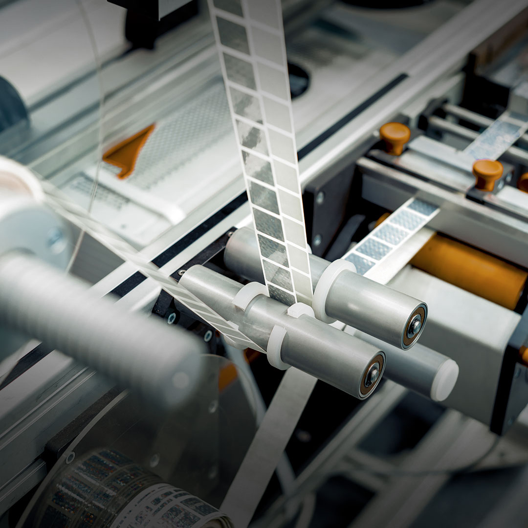 Automated labeling machine applying adhesive labels to transparent film, guided by rollers and mechanical parts in production process.